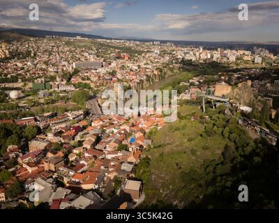 Blick aus der Vogelperspektive auf den Fluss Mtkvari, der sich durch die städtischen Wandteppiche schlängelt, historische Häuser an den Hängen unter einem riesigen Himmel, Tiflis, Georgien. Stockfoto