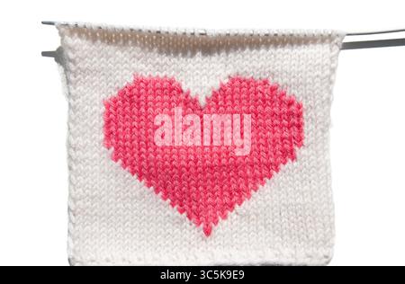 Ein gestricktes rotes Herz auf weißem Hintergrund. Ein rosafarbenes Herz auf weißem Strickmuster auf einer Nadel. Stricknadel mit gestricktem Herzen. Love-Konzept aufbauen Stockfoto