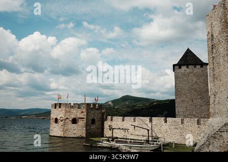 Blick auf eine mittelalterliche Festung Golubac an der Donau mit Türmen, Fahnen und einem vertäuten Holzboot. Historische Stätte in Serbien unter einem dramatischen Himmel. Stockfoto