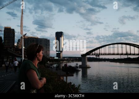Frau genießt die Aussicht bei Sonnenuntergang an der Save in Belgrad, Serbien. Moderne Belgrader Hafengebäude und die alte Brücke bilden einen Kontrast zum alten Stockfoto