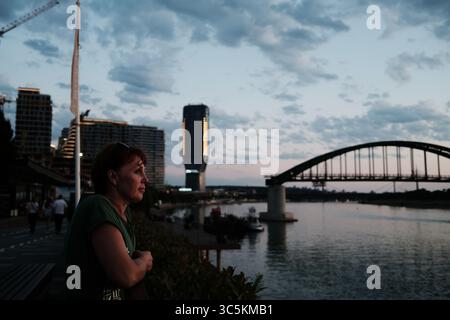 Frau genießt die Aussicht bei Sonnenuntergang an der Save in Belgrad, Serbien. Moderne Belgrader Hafengebäude und die alte Brücke bilden einen Kontrast zum alten Stockfoto