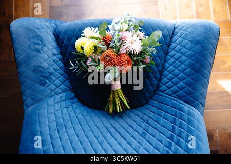 Hochzeitsstrauß mit orangefarbenen Dahlien, rosa und weißen Gerberas, gelben und hellblauen Blumen auf einem blau gesteppten Sessel Stockfoto