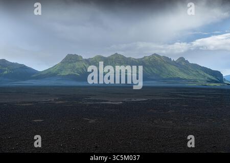Blick auf eine dramatische Landschaft mit dunklem, vulkanischem Gelände, das sich in Richtung majestätischer, grüner Berge unter einem stimmungsvollen Himmel, Kirkjubaejarkla, erstreckt Stockfoto