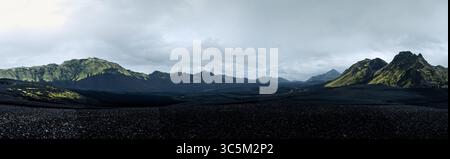 Der Blick auf die schroffe schwarze vulkanische Erde erstreckt sich in Richtung imposanter, grün gefärbter Berge unter einem riesigen, bewölkten Himmel, Langisjor, Skaftárhreppur, Island. Stockfoto