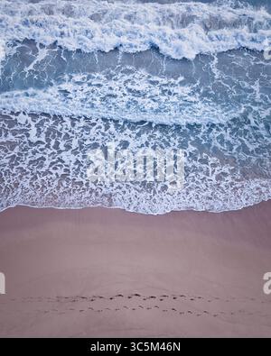 Blick von oben auf sanfte Wellen, die an einem Sandstrand brechen. Im Sand sind Fußspuren im Vordergrund. San Juan, Pr. Stockfoto
