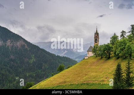 Ein dramatischer Sommersturm zieht über die Dolomiten und tränkt die Landschaft im Regen. Inmitten der nebeligen Gipfel steht Chiesa di Santa Barbara Gracefu Stockfoto