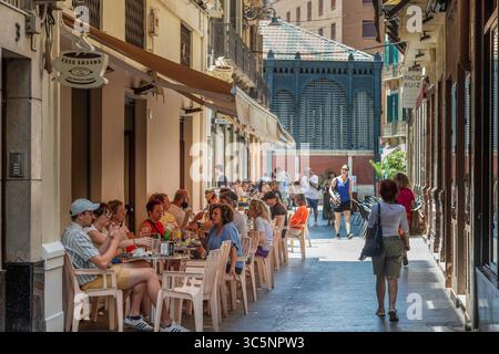 Die Menschen genießen sich auf einer Terrasse in einer engen Gasse im Stadtzentrum von Málaga mit der überdachten Markthalle im Hintergrund. Stockfoto