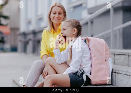 Der junge Schüler isst vor dem Unterricht Apfel mit Rucksack und Mutter in der Nähe und zeigt gesunde Gewohnheiten am Schulmorgen. Stockfoto