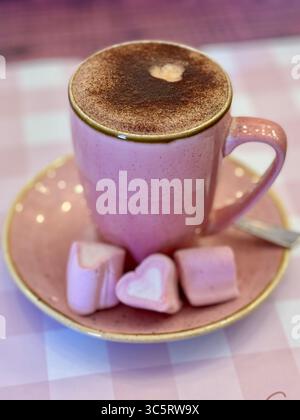 Eine pinkfarbene Kaffeetasse mit einem Cappuccino mit herzförmigem Kakaopulver-Design, serviert mit herzförmigen rosa Marshmallows auf einer passenden Saucette Stockfoto