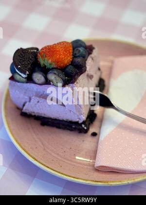 Ein Stück Beeren-Mousse-Kuchen mit Schokoladenkeks-Basis, garniert mit Heidelbeeren, einer Erdbeere und einem Oreo, serviert auf einem rosafarbenen Teller mit einem Herz na Stockfoto