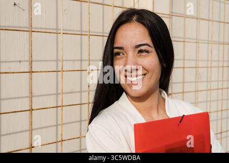 Porträt einer fröhlichen jungen Berufsfrau, die einen roten Ordner hält, der Vertrauen und Erfolg in ihrer Karriere ausstrahlt Stockfoto