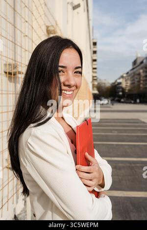 Porträt einer lächelnden jungen Geschäftsfrau aus lateinamerika, die einen roten Ordner hält, während sie in einer städtischen Umgebung steht Stockfoto