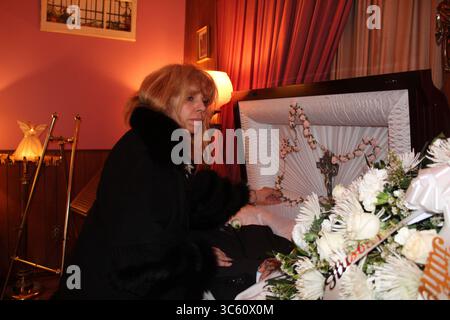 27. April 2020, New York, New York, USA: DORIS V. AMEN legt in einer der schwach beleuchteten Kapellen ihres Bestattungsheims in New York, ihre Gesichtsmaske entfernt, ihre Hand auf den Arm eines verstorbenen älteren Mannes, der in seinem mit weißem Satin gepolsterten Sarg liegt. Das Bestatter-Weibchen sieht eine große Krone weißer Blumen, die den Sarg bedeckt, während es einen großen Rosenkranz berührt. DORIS ist einer der wenigen Mortiker, die die biers offen gelassen haben, um die verstorbenen Personen trotz der Pandemie ihrer Familie zu präsentieren. (Foto: © Marie Le BLE/ZUMA Wire) Stockfoto