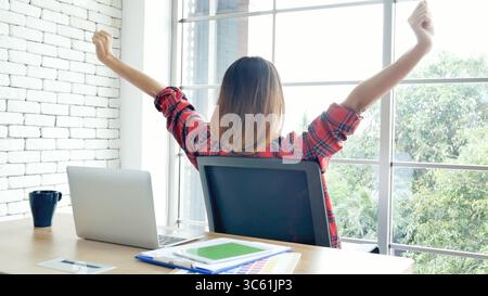 Frau streckt den Arm hoch und sitzt falsch in der Position des Home Office-Schreibtisches. Rückseite einer jungen asiatischen Frau müde von der Arbeit Körper Stress Rücken Schmerz Büro Sy Stockfoto