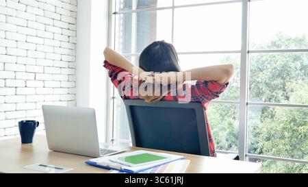 Frau streckt den Arm hoch und sitzt falsch in der Position des Home Office-Schreibtisches. Rückseite einer jungen asiatischen Frau müde von der Arbeit Körper Stress Rücken Schmerz Büro Sy Stockfoto