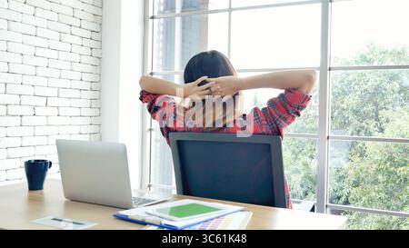 Frau streckt den Arm hoch und sitzt falsch in der Position des Home Office-Schreibtisches. Rückseite einer jungen asiatischen Frau müde von der Arbeit Körper Stress Rücken Schmerz Büro Sy Stockfoto