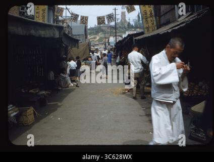 Ein geschäftiger Straßenmarkt in Incheon, Südkorea, 6. Juni 1955. Lokale Händler verkaufen Gemüse, Wurzeln und handgemachte Waren unter Leinwandzelten, während Käufer durch die enge Gasse schlendern. Eine Kirche ist in der Ferne hinter den Marktständen zu sehen. Diese Nachkriegsszene fängt den Alltag während der koreanischen Wiederaufbauzeit ein. Stockfoto