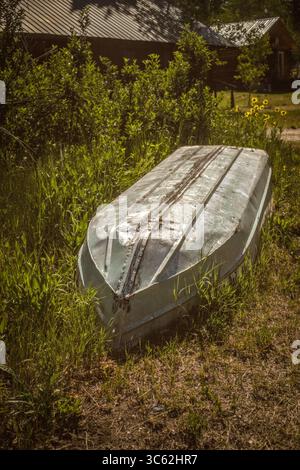 Ein verwittertes Ruderboot aus Aluminium liegt kopfüber in hohem Gras, dessen verblasster Rumpf die Spuren vieler Jahreszeiten trägt. Versteckt neben einer rustikalen Hütte unter dem Stockfoto