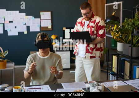Junger Erwachsener kaukasischer Mann mit Virtual-Reality-Headset, interagiert mit digitaler Schnittstelle, während er bärtiger kaukasischer Mann mit Tablet steht und in modernen Büroarbeitsplätzen beobachtet Stockfoto