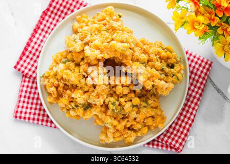 Eine Portion frisch gebratener Corn Fritters (Bakwan Jagung), Golden und knusprig, serviert auf einem weißen Teller mit einem karierten Tuch. Stockfoto