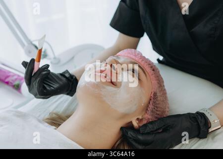Ästhetiker Arzt trägt eine cremige Gesichtsmaske mit einem Pinsel auf das Gesicht der jungen Frau auf Stockfoto