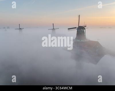 Aus einer dicken Nebeldecke tauchen die Windmühlen aus der Luft auf und schaffen eine traumhafte, surreale Landschaft, Zaandam, Noord-Holland, Niederlande. Stockfoto