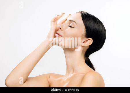 Eine Frau mit klarer Haut berührt sanft ihr Gesicht und hebt eine natürliche Schönheitsroutine hervor, umgeben von sanfter Beleuchtung, die ihren strahlenden Eindruck verstärkt Stockfoto
