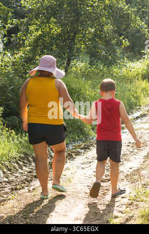 Eine Frau mit Sonnenhut und ein Kind laufen auf einem unbefestigten Weg durch hohes Gras. Sie halten Hände und genießen einen sonnigen Sommertag im Freien. Stockfoto