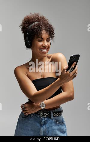 Eine stilvolle Frau mit lockigem Haar trägt Kopfhörer und lächelt, während sie ihr Smartphone benutzt. Stockfoto