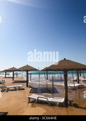 Leerer Strand mit Sonnenschirmen und Wellen, die an sonnigen Tagen Liegen überfluten Stockfoto