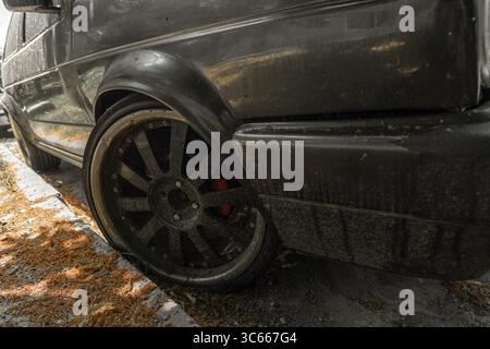 Nahaufnahme eines flachen Hinterrads an einem schmutzigen, verbeulten schwarzen Auto, das auf einer Straße geparkt ist, die mit trockenen Blüten und Trümmern bedeckt ist. Stockfoto