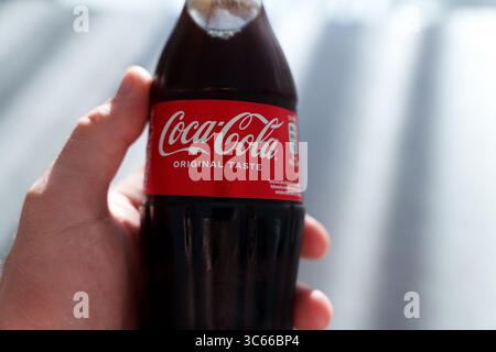 Hand hält eine kalte Coca-Cola Original Geschmacksflasche aus Glas mit rotem Etikett und Kondenswasser, mit Schatten auf einer strukturierten Oberfläche gegossen Stockfoto