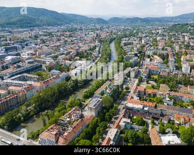Aus der Vogelperspektive auf die Mur, die sich durch die Stadt schlängelt, Gebäude mit roten Dächern im Kontrast zu den grünen Bäumen und den fernen Bergen, Graz, Steiermark, Österreich. Stockfoto