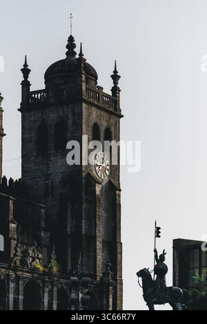 Porto, Portugal - 17. Juni 2022: Blick auf die imposante verwitterte Steinfassade und den Uhrturm der Kathedrale von Porto, hoch neben der Reiterstatue, im sanften Licht getaucht. Stockfoto