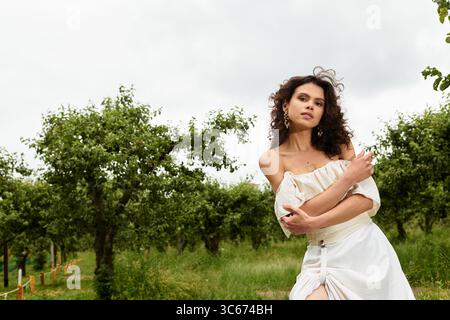 Eine junge Frau in einem weißen Sommerkleid genießt die Natur, ihr lockiges Haar fließt in der Brise. Stockfoto