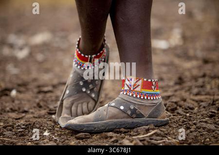 Blick auf lebendige Fußkettchen mit Perlen zieren dunkle Haut über abgenutzten Ledersandalen, die auf dem strukturierten Erdboden stehen, Narok, Narok, Kenia. Stockfoto