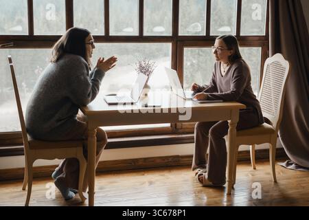 Zwei junge Frauen arbeiten an einem Start-up, arbeiten mit Laptops und diskutieren. Weibliche Geschäftsleute, arbeiten aus der Ferne, hören aufmerksam zu Strategic Stockfoto