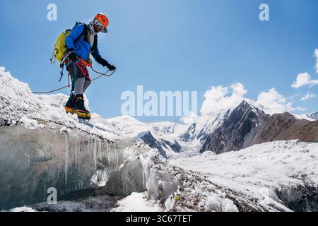 Bergsteiger in pulsierender Ausrüstung hüpft über Gletscherspalten beim Aufstieg des Lenin Gipfels unter klarem Himmel mit schneebedeckten Gipfeln im Hintergrund. Extremer Akt Stockfoto