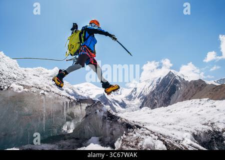 Bergsteiger in pulsierender Ausrüstung hüpft über Gletscherspalten beim Aufstieg des Lenin Gipfels unter klarem Himmel mit schneebedeckten Gipfeln im Hintergrund. Extremer Akt Stockfoto