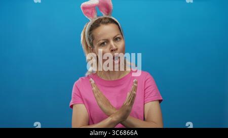Die blonde Frau trägt ein Haarband mit Hasenohren und ein rosafarbenes Hemd kreuzt die Arme im hellen Studio; Ablehnung. Stockfoto