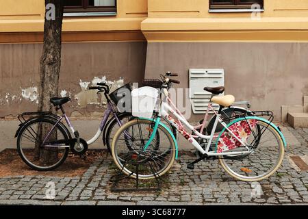 Zwei farbenfrohe Fahrräder parken auf einer kopfsteingepflasterten Straße neben einem Gebäude in einer kleinen serbischen Stadt. Sombor, Serbien - 18. Juli 2025. Stockfoto