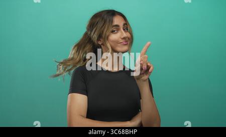 Junge blonde Frau zeigt den Finger in einem grünen Fotostudio nach oben; Selbstvertrauen Verspieltheit Motivation Stärkung. Stockfoto