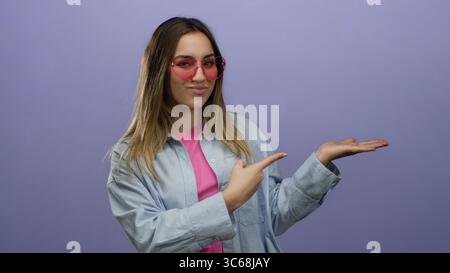 Frau, die eine Geste in einem blauen Hemd mit verspieltem Ausdruck macht, die auf ihre offene Handfläche über einem leuchtend violetten Hintergrund zeigt, mit rosa Sonnenbrillen und Stockfoto