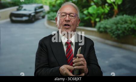 Ein Senior-Geschäftsmann zählt uns im Freien Dollar mit einem Luxusauto im Hintergrund, der Reichtum und Erfolg in einer modernen urbanen Umgebung demonstriert. Stockfoto