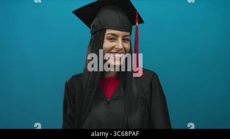 Hispanische Absolvent Frau in Mütze und Kleid, die Stille über isolierte blaue Hintergrundwand symbolisiert Geheimhaltung und Ruhe. Stockfoto