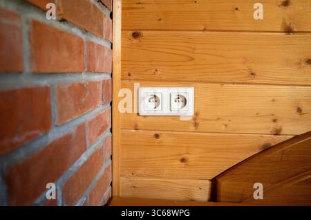Weißer Stecker mit zwei Steckdosen befindet sich an einer Holzwand. Die Mauer ist aus Holz und hat eine gemauerte Umrandung Stockfoto