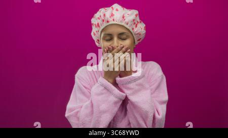 Die junge hispanische Frau im Bademantel mit Duschhaube steht mit überraschtem Ausdruck vor einer isolierten rosa Wand und verkörpert die tägliche Hautpflege-Routine in einem Stockfoto
