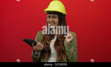Die hispanische Ingenieurin mit gelbem Schutzhelm hält eine Tablette vor einem kräftigen roten Hintergrund und drückt Frustration, Energie und Entschlossenheit aus Stockfoto