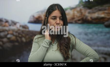 Junge hispanische Frau, die am Strand steht und nachdenklich in Richtung des ruhigen Ozeans unter klarem Himmel blickt und eine natürliche kontemplative Stimmung zeigt Stockfoto