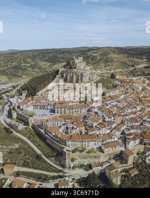 Blick aus der Vogelperspektive auf die alten Steinmauern umgeben eine Stadt mit roten Ziegeldächern, gekrönt von einer majestätischen Burg vor der Kulisse der sanften grünen Hügel., Morella, Valencianische Gemeinde, Spanien. Stockfoto